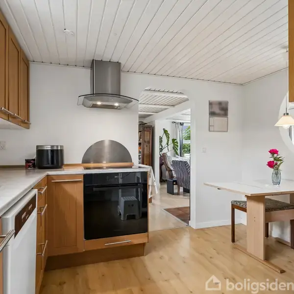 Køkken med træskabe og hvid bordplade inkluderer ovn, komfur og emhætte. Det er forbundet til stue gennem en bueåbning. Et lille spisebord med blomster står ved siden af.