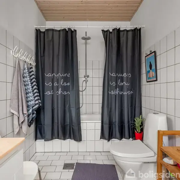 Et badeværelse med et brusebad og toilet. Mørke gardiner over badekarret har tekst: "Happiness is a long hot shower." Lyse vægge, fliser og træloft. Håndklæder hænger ved siden af bruseren. En lille plante står ved toilettet, og der er en stige-hylde ved siden af vasken.