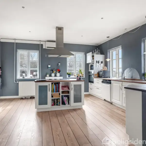 Et moderne køkken med hvidt interiør, træbordplader og en central ø med indbygget bogreol. Rummet er lyst med flere vinduer, hængende lamper og grå vægge.
