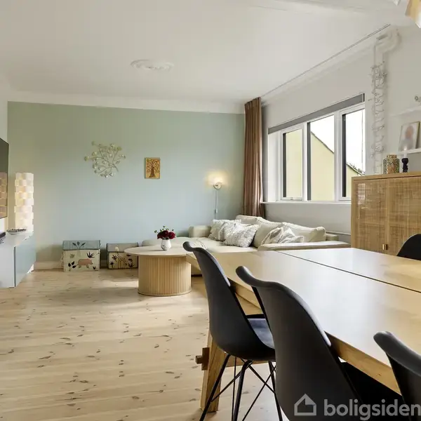 Spisebord med sorte stole står i lys stue med trægulve og lyseblå vægge. I baggrunden er der en lys sofa ved siden af et vindue.