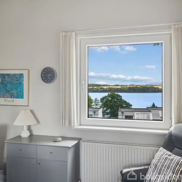 Et rum med en behagelig lænestol, hvor en lampe og en skænk står ved væggen. Vinduet viser en udsigt over en sø med træer i baggrunden.