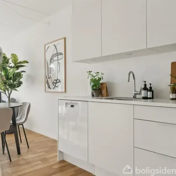 Et moderne køkken med hvide skabe og en køkkenvask. Bordpladen har planter og sæbedispensere. Spisebord med stole og en lys pendellampe i baggrunden. Sollys skaber en lys atmosfære.