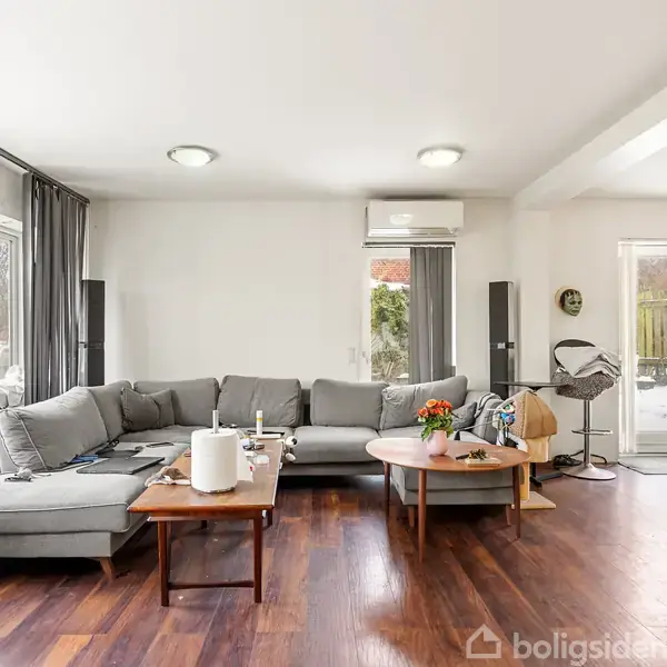 En grå hjørnesofa står placeret i en moderne stue med trægulve. På sofabordet ligger en skål med blomster og papirruller. Rummet har lyse vægge og vinduer mod venstre.