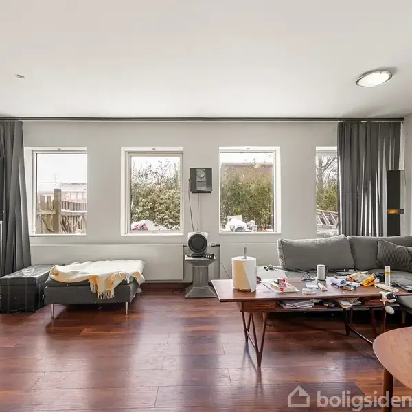 Stue med grå hjørnesofa og sofabord fyldt med diverse genstande. Store vinduer lukker lys ind, mens et gråt skabsarrangement og flyttekasser fra jem & fix ses i baggrunden.