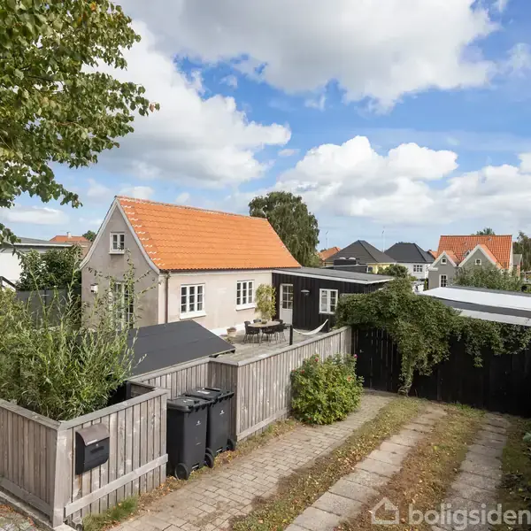 Hus med orange tag står stille i en have med hæk og træhegn. Omkringliggende huse fylder nabolaget under en skyet, blå himmel. To skraldespande ses ved indgangen.