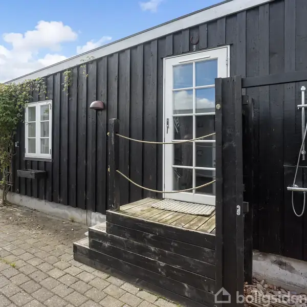 Sort træhus med vinduer og dør, omgiven af brostensbelagt terrasse. En udendørs bruser er monteret på væggen. Planter gror omkring huset. Himlen er klar med få skyer.