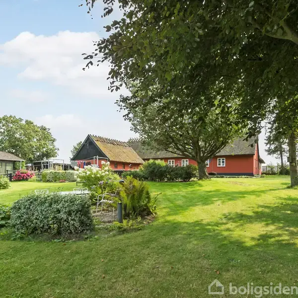Et rødt hus med stråtækt tag står stille i en grøn have omgivet af store træer. En terrasse med møbler er også synlig i baggrunden.