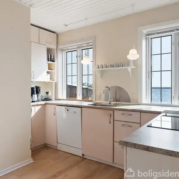Et lyst køkken med indbyggede skabe og hvidevarer, herunder opvaskemaskine og kogeplader. Store vinduer giver udsigt til vandet udenfor, mens to lamper hænger over arbejdsområdet.