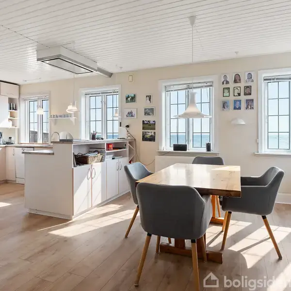 Et lyst køkken med en spisebord og fire grå stole. Rummet har lyse trægulve og hvide lofter, og væggene er dekoreret med billeder. Store vinduer slipper masser af lys ind.