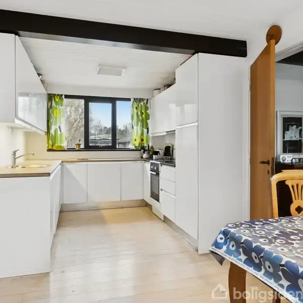 Køkken med hvide skabe og indbygget komfur, bord ved siden med blåmønstret dug, en stol, og vindue med grønne gardiner i baggrunden. Bordlampe hænger over bordet.