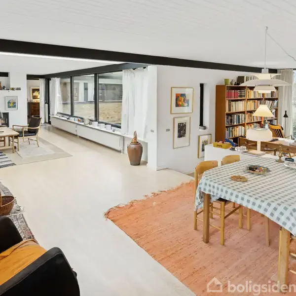 Stue med spisebord dækket med ternet dug, omgivet af træstole. Sofaområde ses til venstre. Hylder fyldt med bøger står op ad vinduerne. Store vinduer giver udsigt til have.