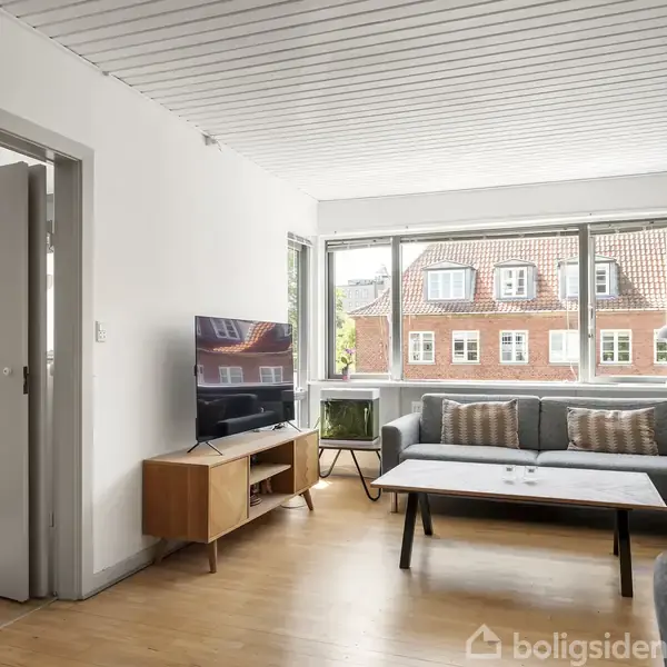 Stue med gråt hjørnesofa og sofabord, samt fladskærms-TV på et træbord. Møblerne er placeret foran store vinduer, der giver udsigt til andre bygninger med røde mursten.