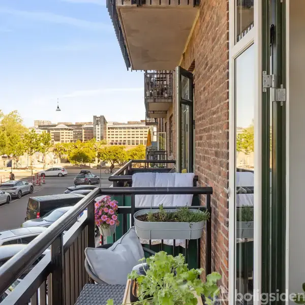 En altan rummer blomsterkasser med lyserøde blomster og krydderurter. Mod en byvej med parkerede biler og moderne bygninger ses grønne træer og blå himmel i baggrunden.