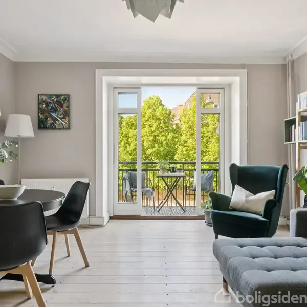 En stue med et spisebord og stole til venstre, en grøn lænestol foran reoler til højre, stor fransk altandør åbner til en balkon omgivet af grønne træer i baggrunden.