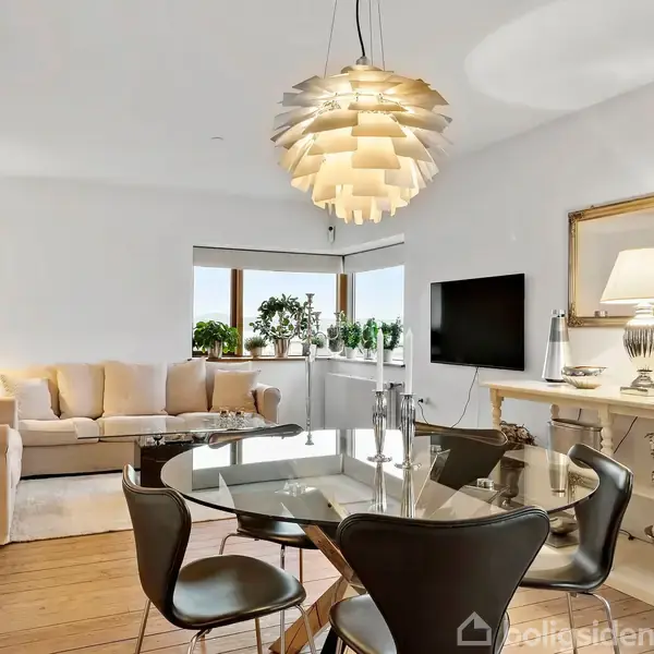 En moderne stue med en rund glasbord omgivet af stole. Over bordet hænger en kunstnerisk lampe. I baggrunden står en beige sofa ved siden af planter ved et vindue. En stor spejlramme og fladskærm pryder væggen.