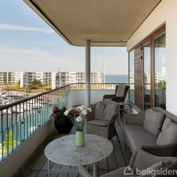 En balkon møbleret med grå sofaer og et rundt bord står stille. Udsigt over en marina med fortøjede både. Højhuse ses i baggrunden ved vandet.