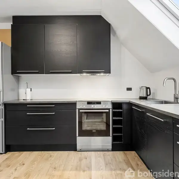 Et moderne køkken med sorte skabe udstyret med en ovn, emhætte og køleskab. En stor bordplade med vask og elkedel står ved siden af et skråvindue, omgivet af trægulv.