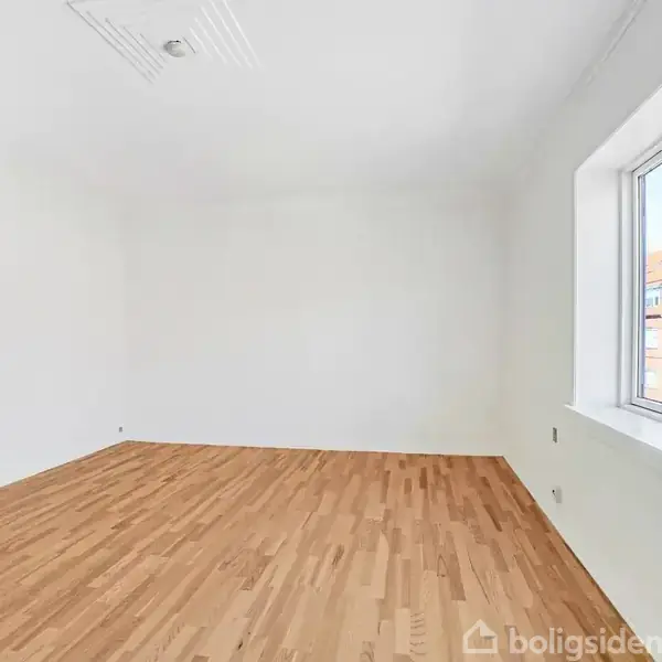 Et tomt rum med trægulv, ingen møbler, store vinduer til højre, der slipper lys ind. Væggene er hvide, og der er udsigt til røde tage udenfor.