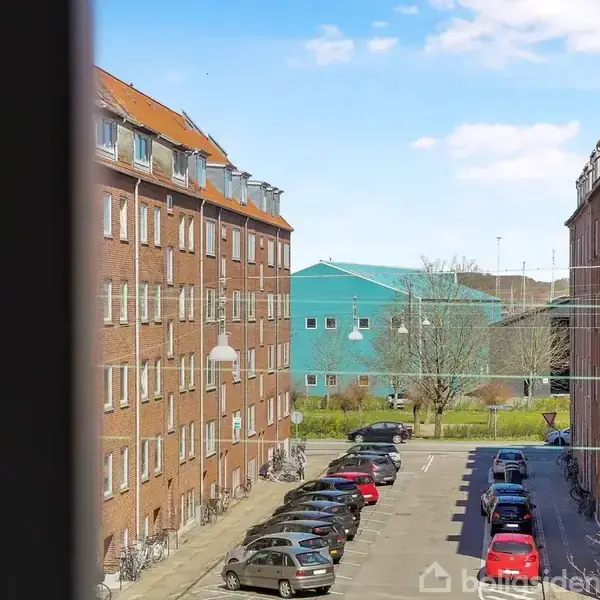 Røde murstensbygninger står på hver side af en parkeringsplads med biler, mens en blå bygning og træer ses i baggrunden under en klar, blå himmel.