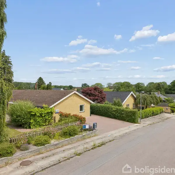 En gul etplanshus med have ligger langs en stille vej i et villakvarter. Der er buske og træer omkring huset, og få biler er parkeret langs vejen.