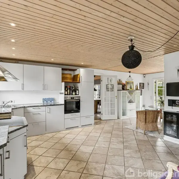 Et moderne køkken med hvide skabe og grå marmorbordplader. En stor rund lampe hænger fra det træloftede loft. I baggrunden ses en spiseplads og en terrasse.