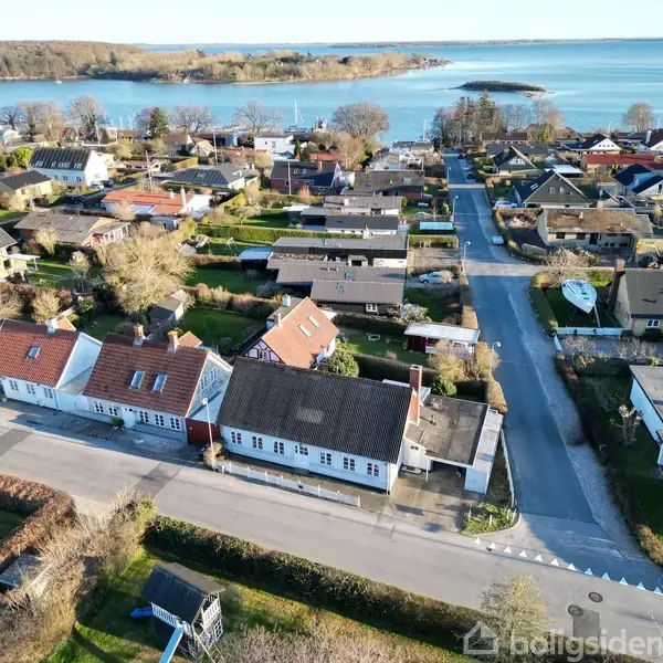 Rækkehus med røde og hvide tage ses fra luften i en kystby. Veje og andre huse omgiver dem, med en fjord og skov i baggrunden.