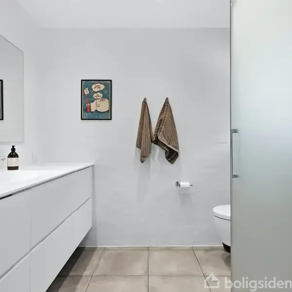 Et hvidt badeværelse med en moderne dobbeltvask og spejl til venstre. To håndklæder hænger på væggen over en toiletpapirrulle. To billeder hænger ved siden af håndklæderne.