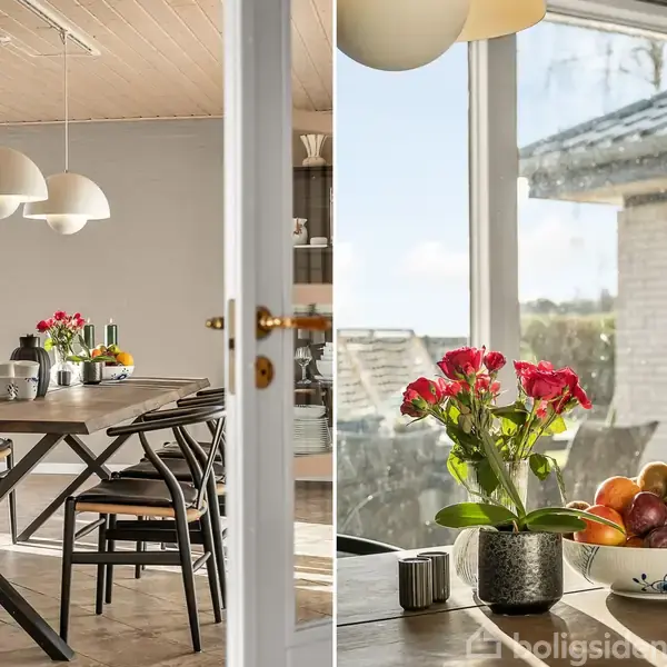 Et spisebord med sorte stole står i en lys spisestue. På bordet er der en vase med røde blomster og en skål med frugt. Store vinduer giver udsigt til huset.