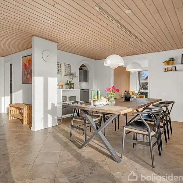Et stort spisebord med sorte stole omkring står i en rummelig spisestue. To hvide lamper hænger over bordet. Omgivelserne inkluderer hvide vægge, flisegulv og en træloft.