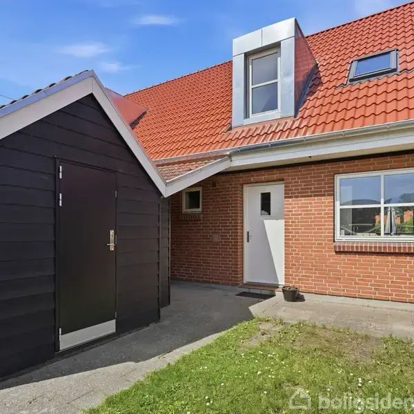 Rødt murstenshus med rødt tag og kvist, beliggende i en have. Til venstre ses en sort træbygning. Foran huset er en græsplæne og en grå gangsti.
