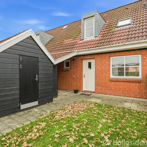 Et rødt murstenshus med rødt tegltag og sort udhus. Huset har en hvid dør og et vindue. Det står i en have med en flisebelagt sti, omkranset af hæk.