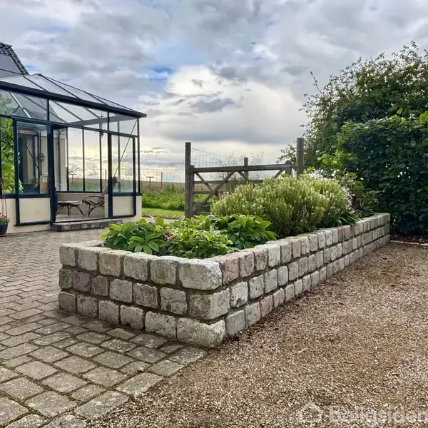 Drivhus stående på en brostensbelagt gårdsplads ved siden af en lav plantekasse i mursten med grønne planter. I baggrunden er der en træport og en tæt busk.