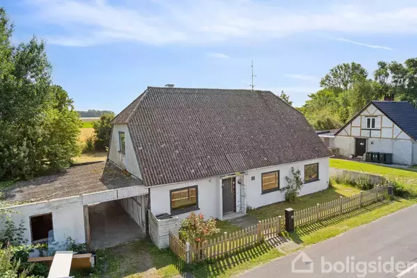Et hvidmalet hus med skråt tag står i en landlig vej omgivet af marker og træer. Der er en carport og et lille hegn i forhaven.