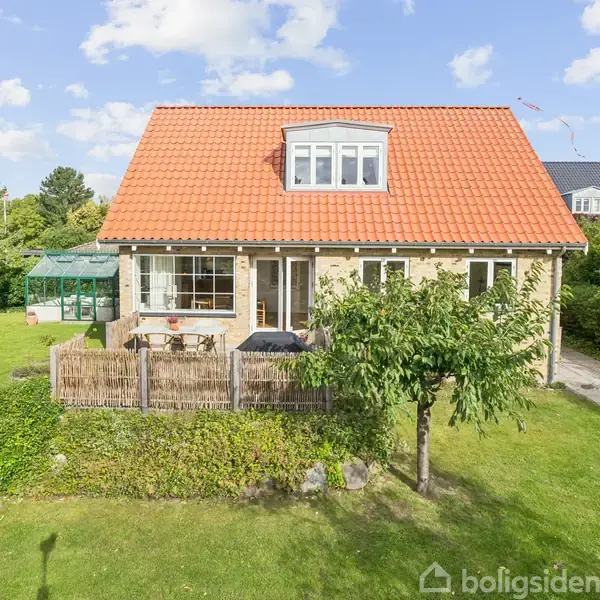 Et gult hus med en rød tag beliggende i en grøn have med træer og en terrasse. Et drivhus står i baggrunden. Skyer og blå himmel ses over huset.