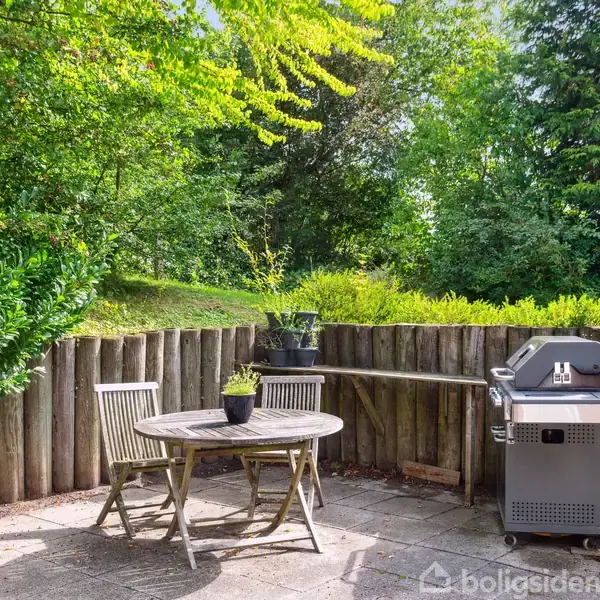 Et rundt bord med to stole står stille på en terrasse omgivet af træstolper og frodig grøn vegetation. En grill er placeret til højre i uderummet.