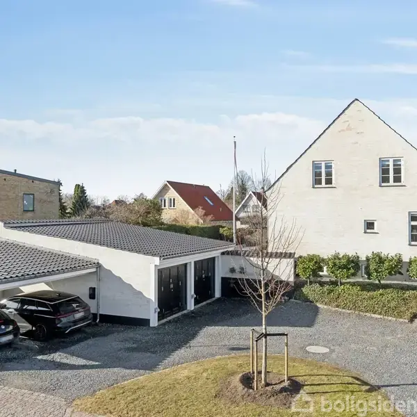 Huset står stillet i et villakvarter med en hvid facade og sort tag. Til venstre holder to biler under en garage, omgivet af en grusbelagt indkørsel og små buske.
