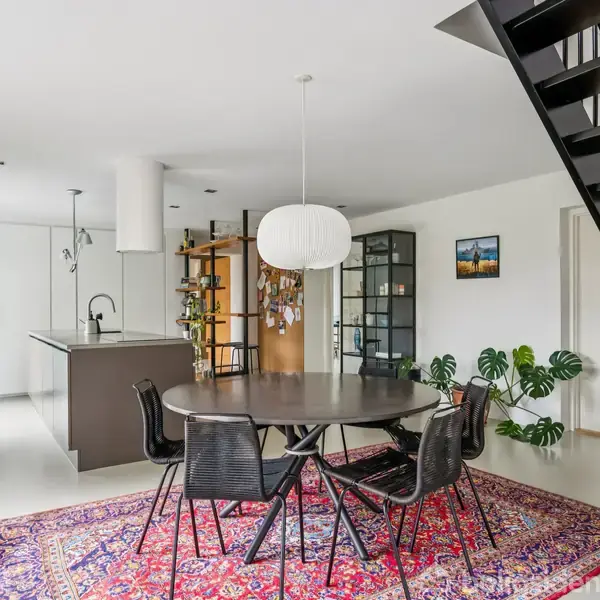 Rundt spisebord med stole står på et farverigt tæppe. Moderne køkken i baggrunden med hvidt interiør og øenhed. Åben trappe til højre og planter rundt omkring.