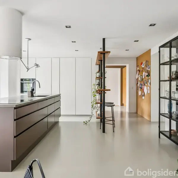 Køkken med køkkenø, stoler op mod barhæng, og åben reol fyldt med glas og bøger. Grøn væg til venstre og opslagstavle med fotos i baggrunden.
