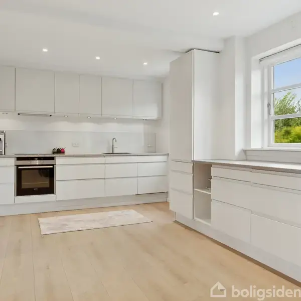 Et moderne køkken har hvide skabe og apparater. Skabene er monteret over en ovn og en vask. Et vindue giver udsigt til grønne træer, og gulvet er af lyst træ.