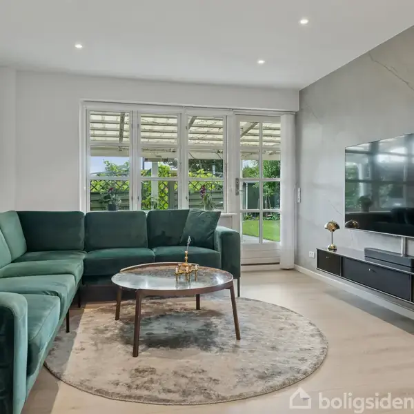 Grøn sofa står ved stuebord på rund tæppe, foran en stor fladskærms-tv monteret på væggen. Store vinduer i baggrunden giver udsigt til en have.