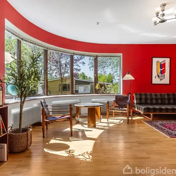 Stue med rød væg, sofa, og rundt sofabord i træ, oplyst af naturligt lys fra et stort rundt vindue. På gulvet ligger et tæppe, og der står en gulvlampe ved siden af sofaen. Reol med bøger og en plante i kurv står ved væggen.