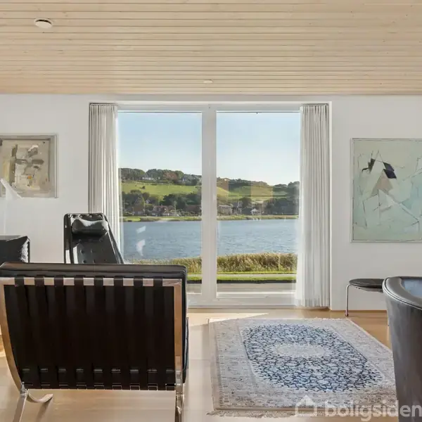 En stue med moderne møbler som sofaer og lænestole, orienteret mod store vinduer med udsigt til en sø og grønne bakker udenfor. Vægge prydet med kunstværker.