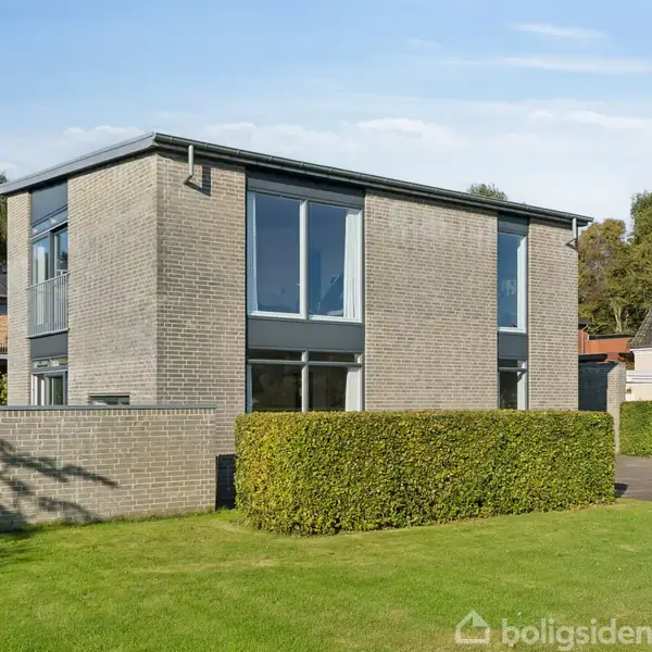 Et moderne, to-etagers murstenshus med store vinduer står i en velplejet have omkranset af hække. Det ligger i et roligt boligområde med nabohuse i baggrunden.