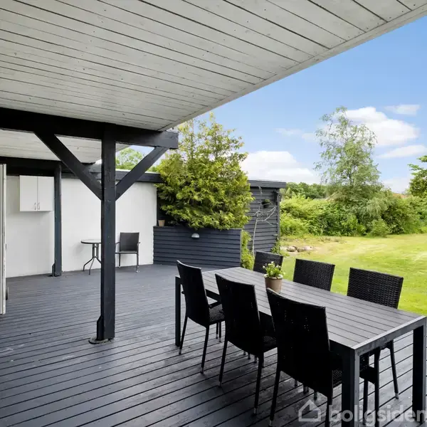 Overdækket terrasse med sort spisebord og stole, grill i hjørnet. Terrassen har udsigt til en velplejet græsplæne omgivet af buske og træer under en klar blå himmel.
