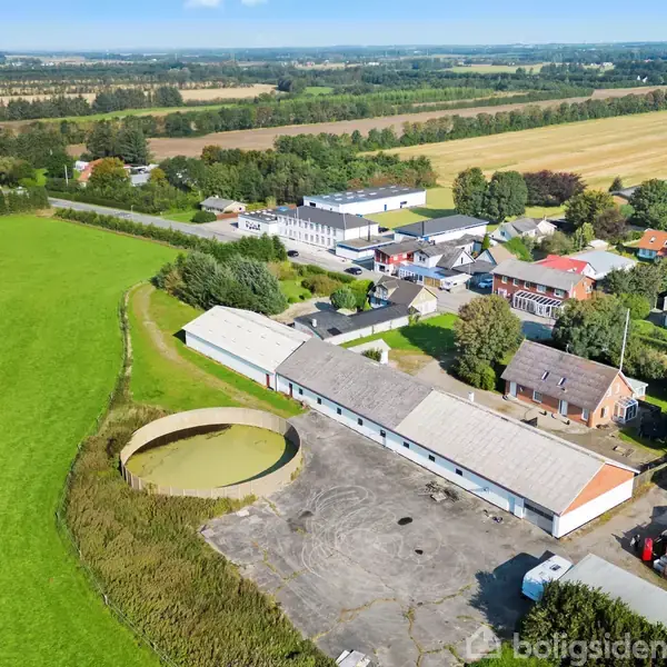 Bygninger og silo stille ved en stor gårdsplads, omgivet af grønne marker og landskabelige områder. Små huse og træer ses i nærheden af det maleriske landsbymiljø.