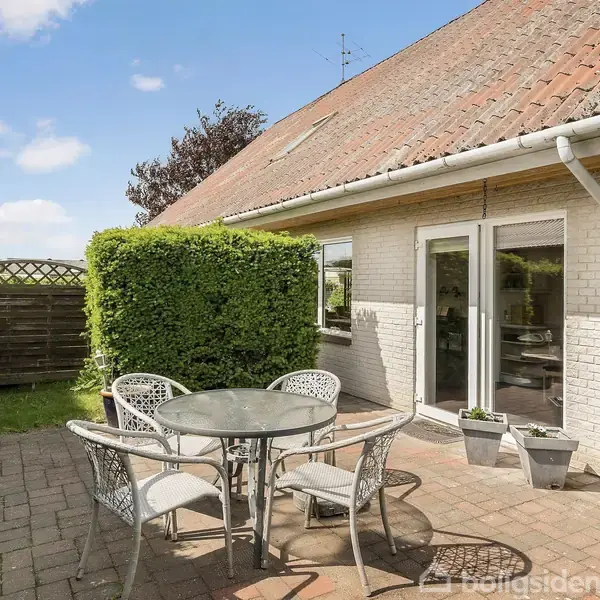 En rund metalbord med fire stole står på en flisebelagt terrasse. I baggrunden ses en hvidmalet husmur med store vinduer og en grøn hæk.