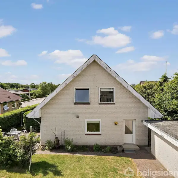 Hus med spidstag, to vinduer og hvid murstensfacade står stille i en velplejet have med græsplæne, omgivet af buske og træer under en klar blå himmel.