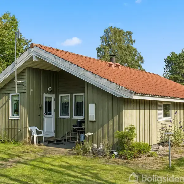 Et gråt træhus med rødt tag står i en have omgivet af grønne træer. En stol og et bord er placeret ved indgangen. Himlen er blå og klar.