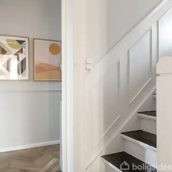 En hvidmalet trappe stiger, omgivet af en lys væg med to abstrakte kunstværker. Mønstre og cirkelmotiver pryder væggen, hvilket skaber en harmonisk og moderne atmosfære.