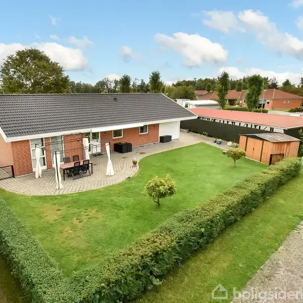 Huset er en moderne, enkeltstående bygning med rødt murværk og sort tag. Terrasse med havemøbler foran, omgivet af velplejet have med hække. Nabolag med flere lignende huse ses omkring.